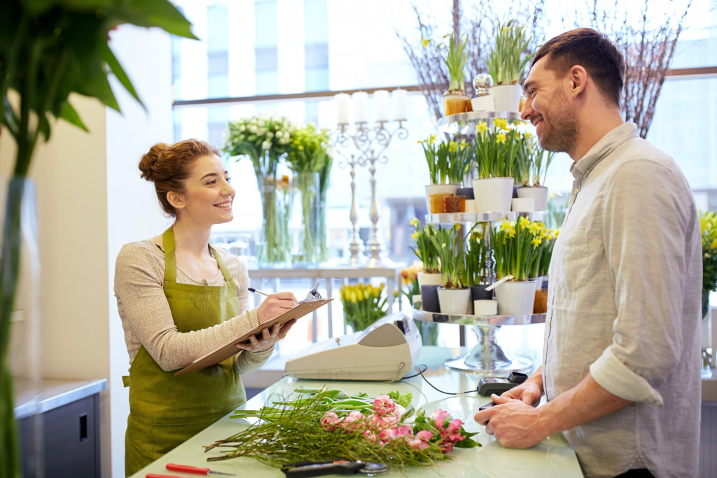 A florist smiles while holding a pen and clipboard talking to a customer at the flower shop